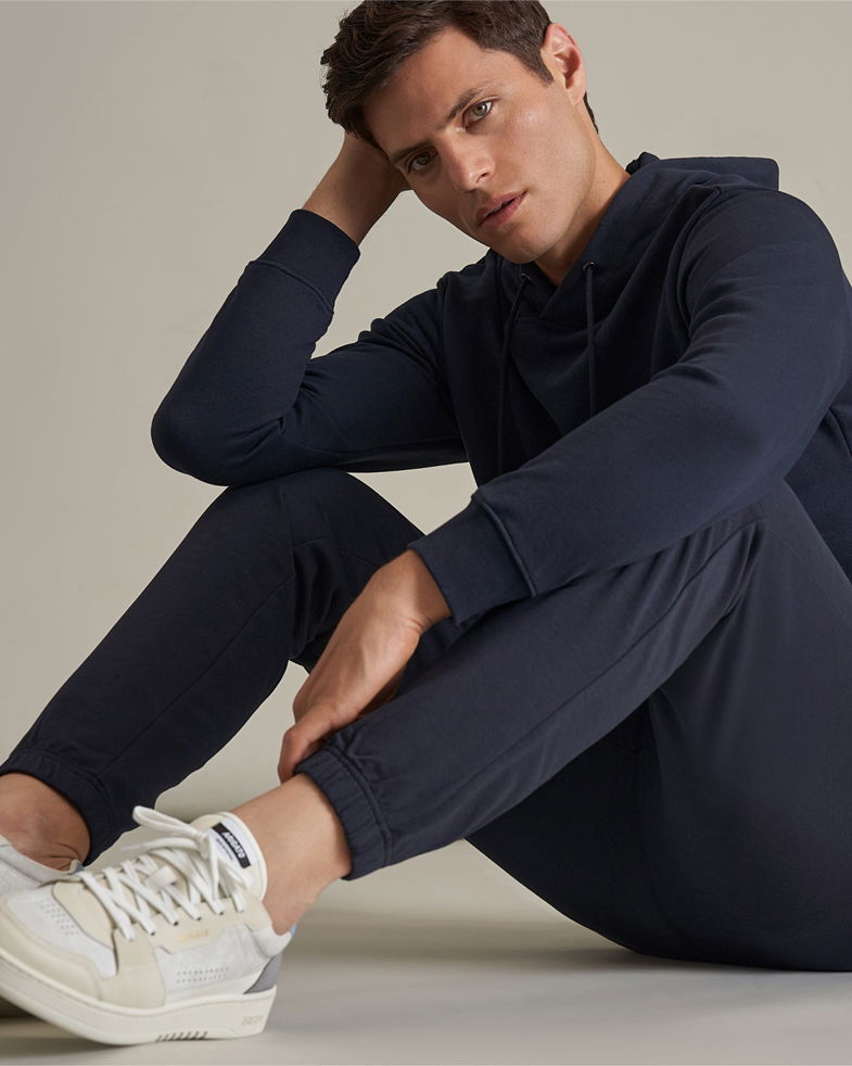 Mann sitzt entspannt, trägt dunkelblauen Hoodie, Joggpants und hellgrau-beige Sneaker auf hellem Hintergrund.