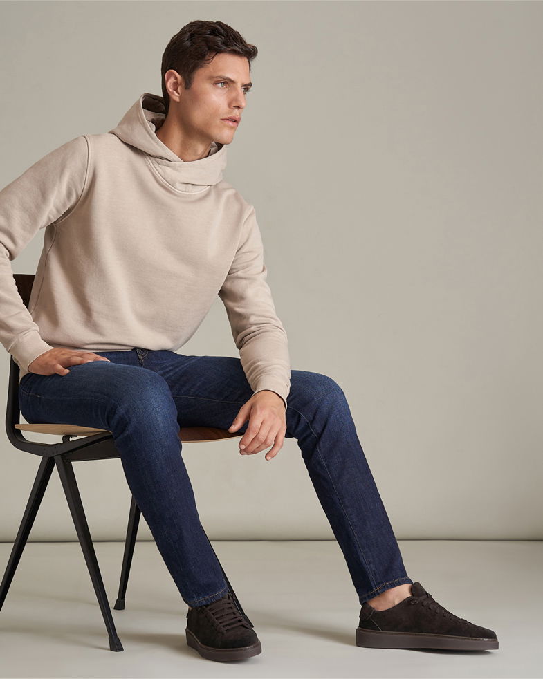 Junger Mann sitzt auf Stuhl, trägt beigen Hoodie, dunkelblaue Slim-Fit-Jeans und dunkelbraune Sneaker, neutraler Hintergrund.