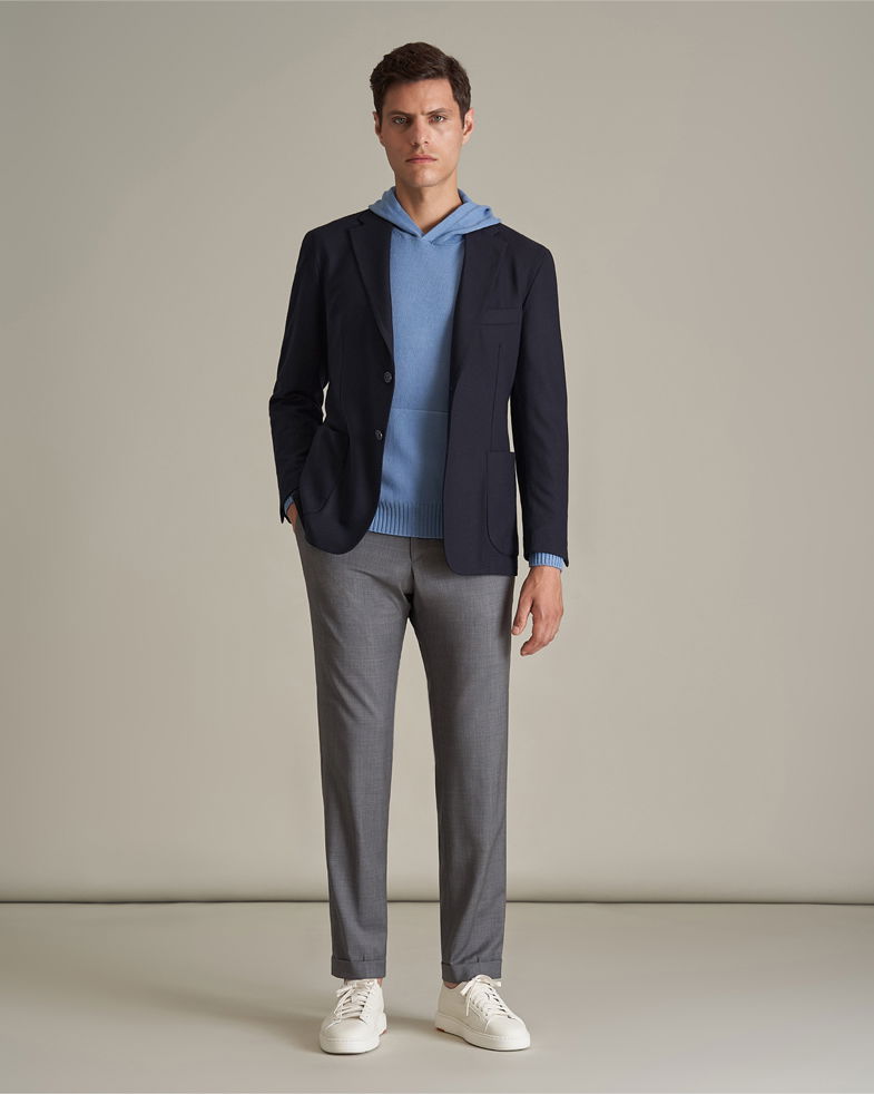 Mann trägt rauchblauen Hoodie, navy Sakko, dunkelgraue Hose und weiße Sneaker vor beigem Hintergrund.