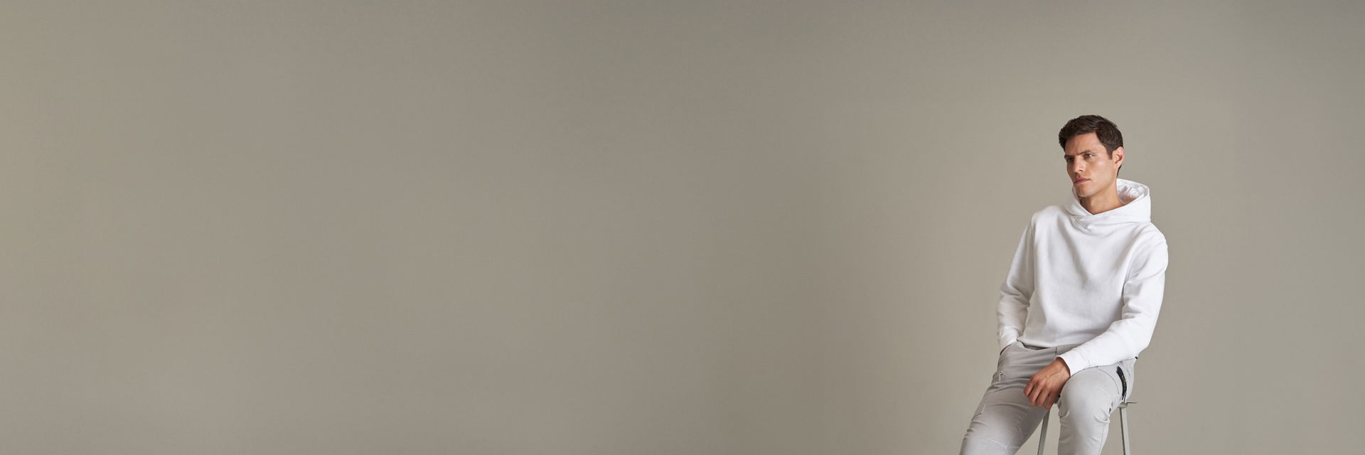 Mann sitzt auf Hocker, trägt weißen Hoodie und hellgraue Cargohose, neutraler Hintergrund, nachdenklicher Gesichtsausdruck. Mann sitzt auf Hocker, trägt weißen Hoodie und hellgraue Cargohose, neutraler Hintergrund, nachdenklicher Gesichtsausdruck.