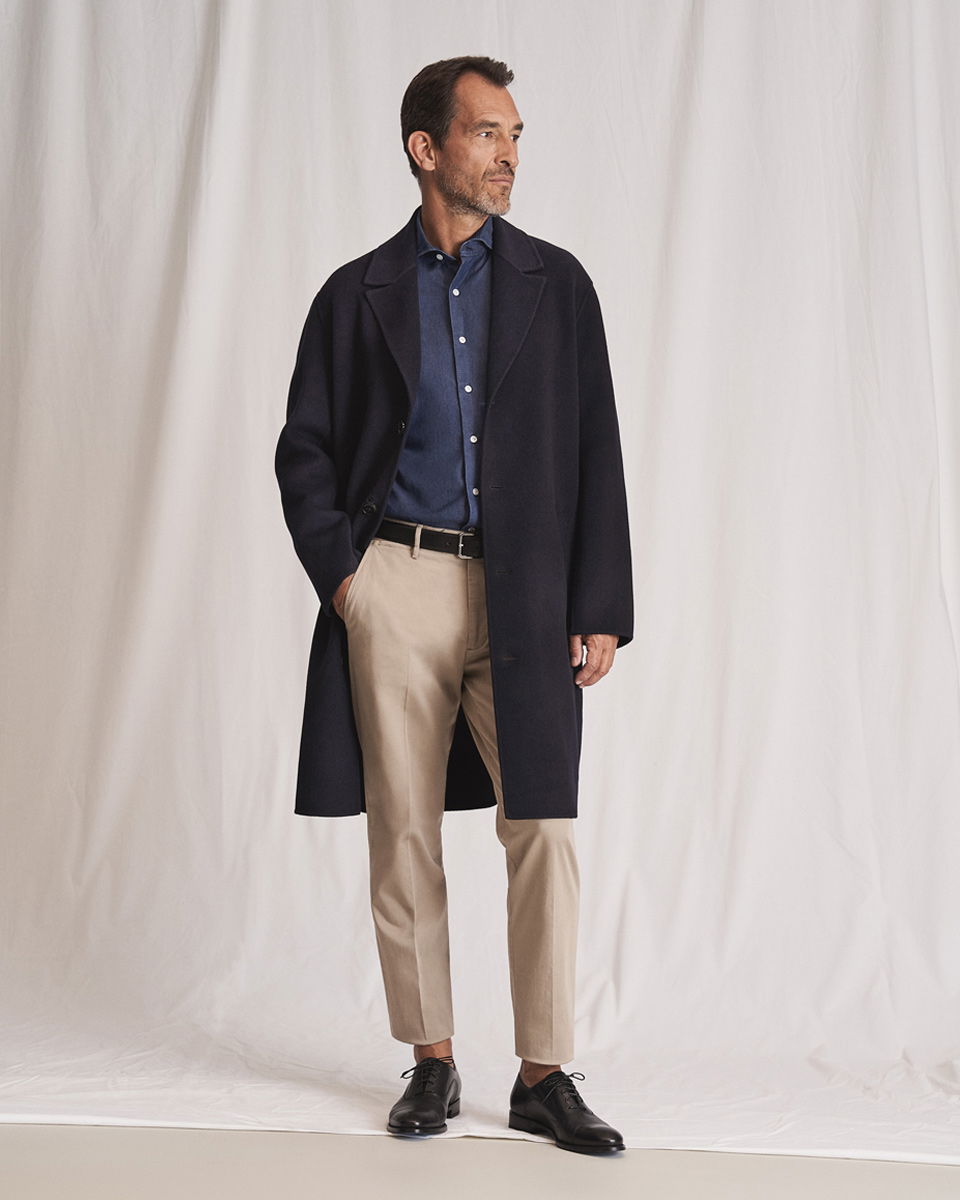 Mann trägt schwarzen Mantel, dunkelblaues Hemd, beige Hose, schwarzen Gürtel und schwarze Schnürschuhe vor hellem Hintergrund.