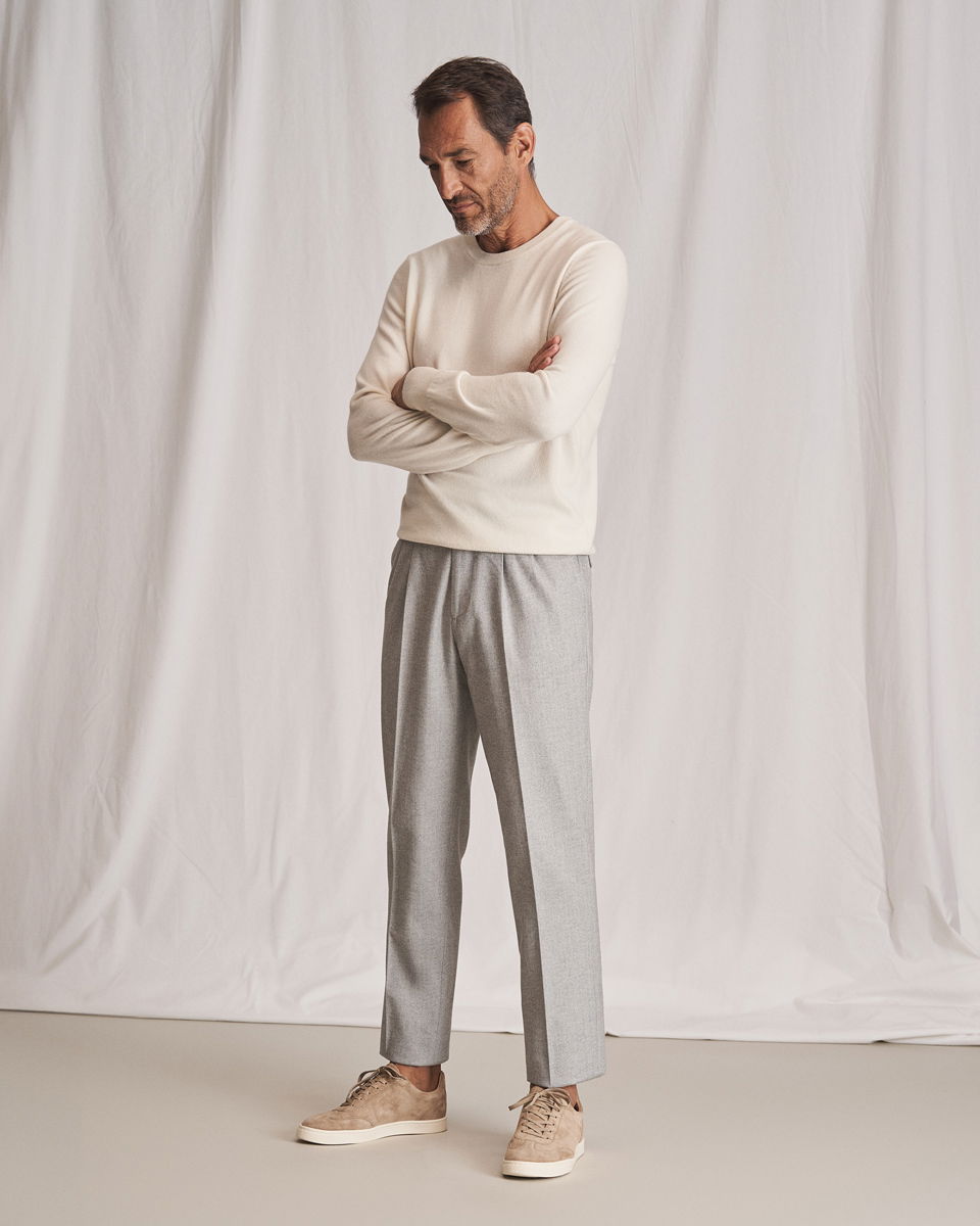 Mann in cremefarbenem Rundhalspullover, hellgrauer Hose und beigen Sneakern steht vor weißem Hintergrund mit verschränkten Armen.