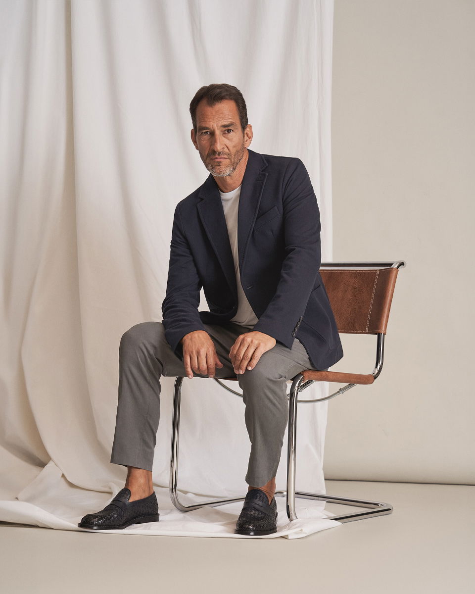 Mann sitzt auf Stuhl, trägt dunkelblaues Sakko, weißen Pullover, graue Hose und schwarze Loafer mit Flechtoptik.