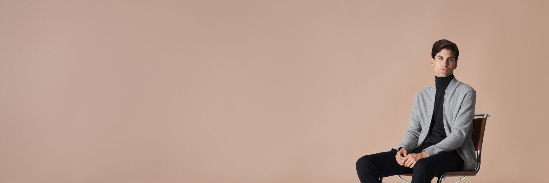 Mann sitzt auf Stuhl vor beigem Hintergrund, trägt graue Strickblouson, schwarzen Rollkragenpullover, schwarze Hose und Chelsea Boots. Mann sitzt auf Stuhl vor beigem Hintergrund, trägt graue Strickblouson, schwarzen Rollkragenpullover, schwarze Hose und Chelsea Boots.