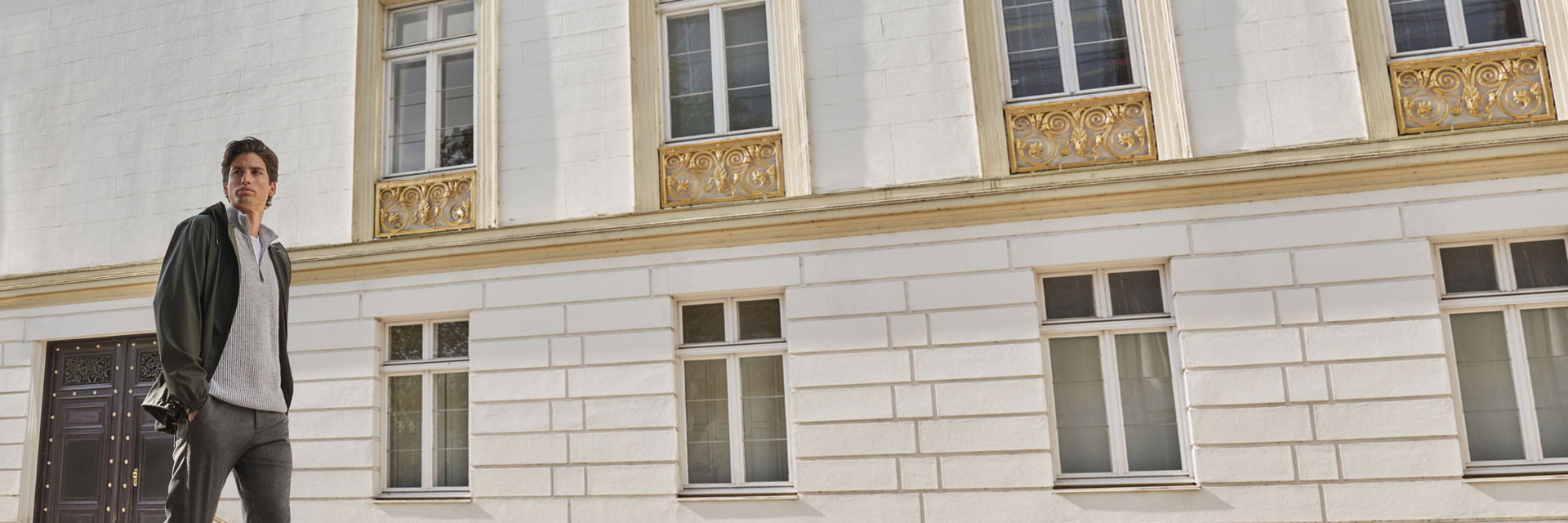 Mann trägt hellgrauen Troyer, weißes T-Shirt, olivgrüne Jacke und dunkelgraue Wollhose vor weißem Gebäude mit goldverzierten Fenstern. Mann trägt hellgrauen Troyer, weißes T-Shirt, olivgrüne Jacke und dunkelgraue Wollhose vor weißem Gebäude mit goldverzierten Fenstern.
