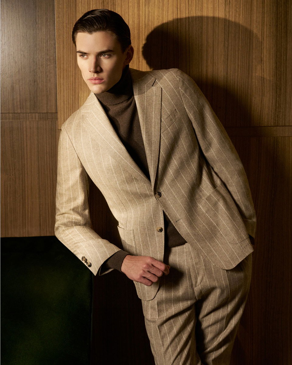Junger Mann lehnt an Holzpaneelen, trägt beigen Nadelstreifenanzug und braunen Rollkragenpullover, schaut nach links. Junger Mann lehnt an Holzpaneelen, trägt beigen Nadelstreifenanzug und braunen Rollkragenpullover, schaut nach links.