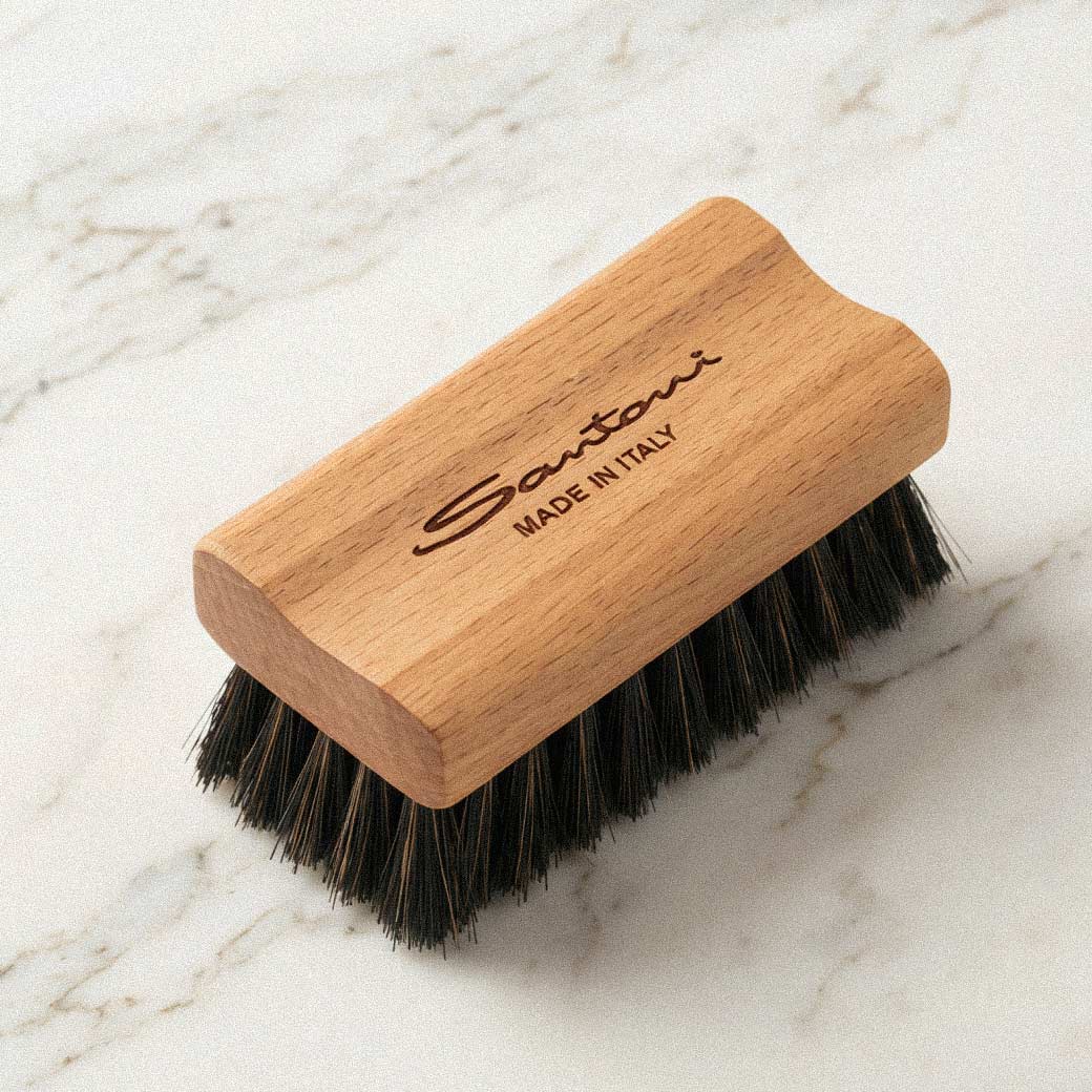 Holzbürste mit schwarzen Borsten, Gravur 'Santoni Made in Italy' auf Holzgriff, liegt auf marmoriertem hellen Untergrund. Holzbürste mit schwarzen Borsten, Gravur 'Santoni Made in Italy' auf Holzgriff, liegt auf marmoriertem hellen Untergrund.