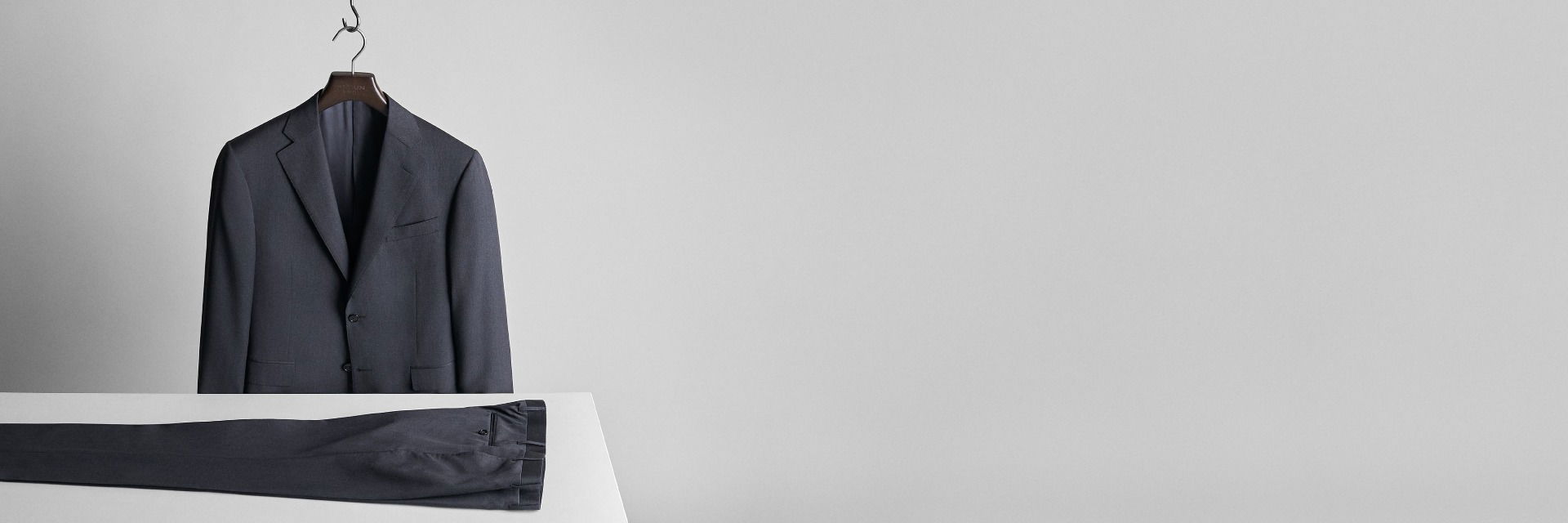 Anthrazitfarbenes Sakko an Bügel hängend, davor passende Hose liegend auf weißem Tisch vor grauem Hintergrund. Anthrazitfarbenes Sakko an Bügel hängend, davor passende Hose liegend auf weißem Tisch vor grauem Hintergrund.