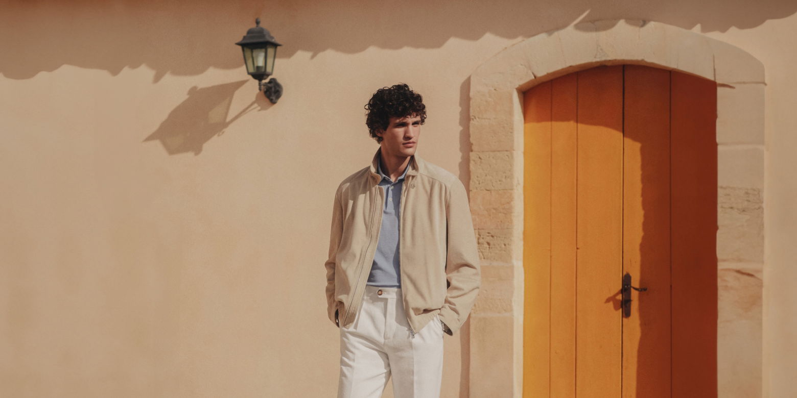 Mann mit lockigem Haar trägt beige Velourslederjacke, hellblaues Poloshirt und weiße Hose vor orangefarbener Tür. Mann mit lockigem Haar trägt beige Velourslederjacke, hellblaues Poloshirt und weiße Hose vor orangefarbener Tür.