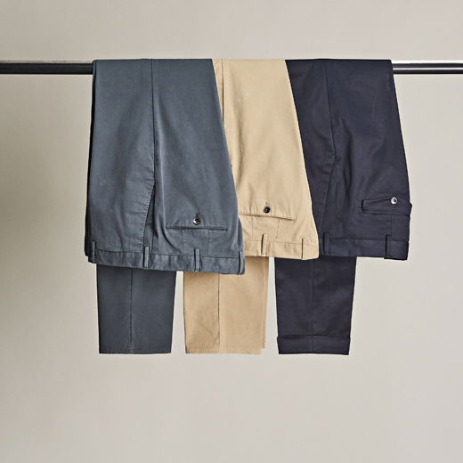 Drei Herren-Chinohosen in Grau, Beige und Navy, ordentlich über eine Kleiderstange hängend vor hellem Hintergrund. Drei Herren-Chinohosen in Grau, Beige und Navy, ordentlich über eine Kleiderstange hängend vor hellem Hintergrund.