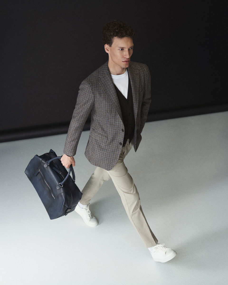 Mann läuft, trägt kariertes Sakko, schwarzen Cardigan, weißes Shirt, beige Hose, weiße Sneaker, hält navyblaue Tasche in der Hand.