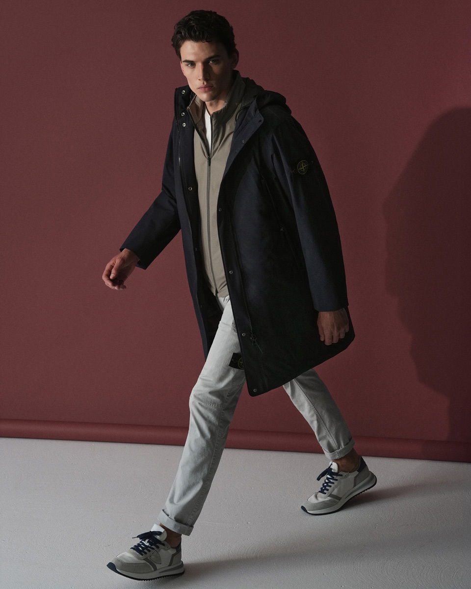 Mann läuft vor rotem Hintergrund mit hellgrauer Cargohose, beiger Sweatjacke, blauem Parka und grauen Sneakers.