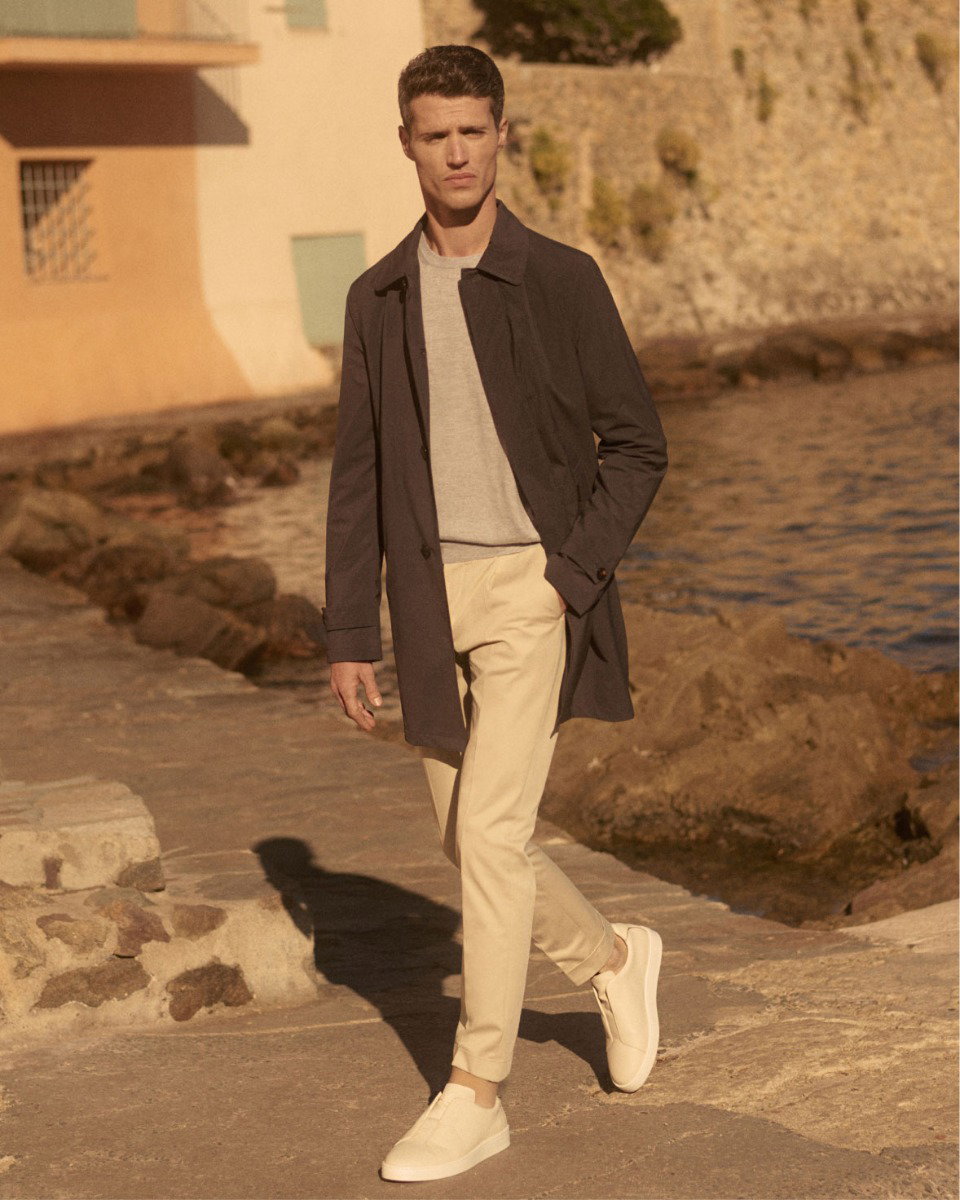 Mann läuft am Wasser entlang, trägt dunkelgrauen Kurzmantel, hellbeige Chinohose und weiße Sneaker.