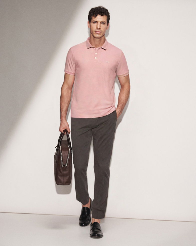 Mann läuft, trägt rosa Poloshirt, dunkelgraue Hose, schwarze Loafer und hält braune Ledertasche in der Hand.