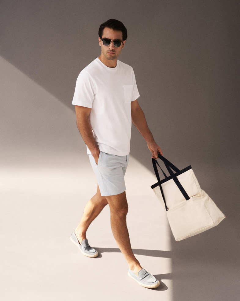 Mann mit weißem T-Shirt, hellblauen Shorts, grauen Espadrilles und cremefarbener Tasche läuft vor grauem Hintergrund. Mann mit weißem T-Shirt, hellblauen Shorts, grauen Espadrilles und cremefarbener Tasche läuft vor grauem Hintergrund.