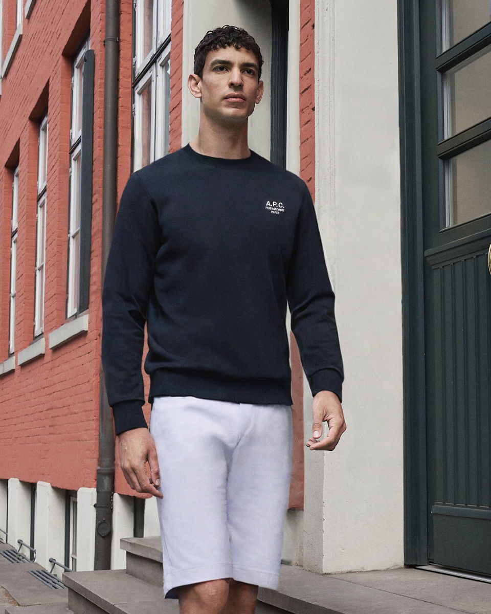 Mann läuft vor rotem Backsteingebäude, trägt schwarzen Pullover mit Logo und weiße kurze Hose.