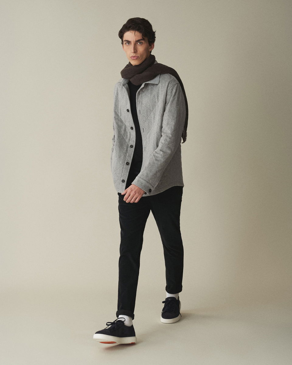 Junger Mann läuft, trägt graues gesteppte Overhirt, schwarzen Pullover, schwarze Hose, dunklen Schal und schwarze Sneaker.