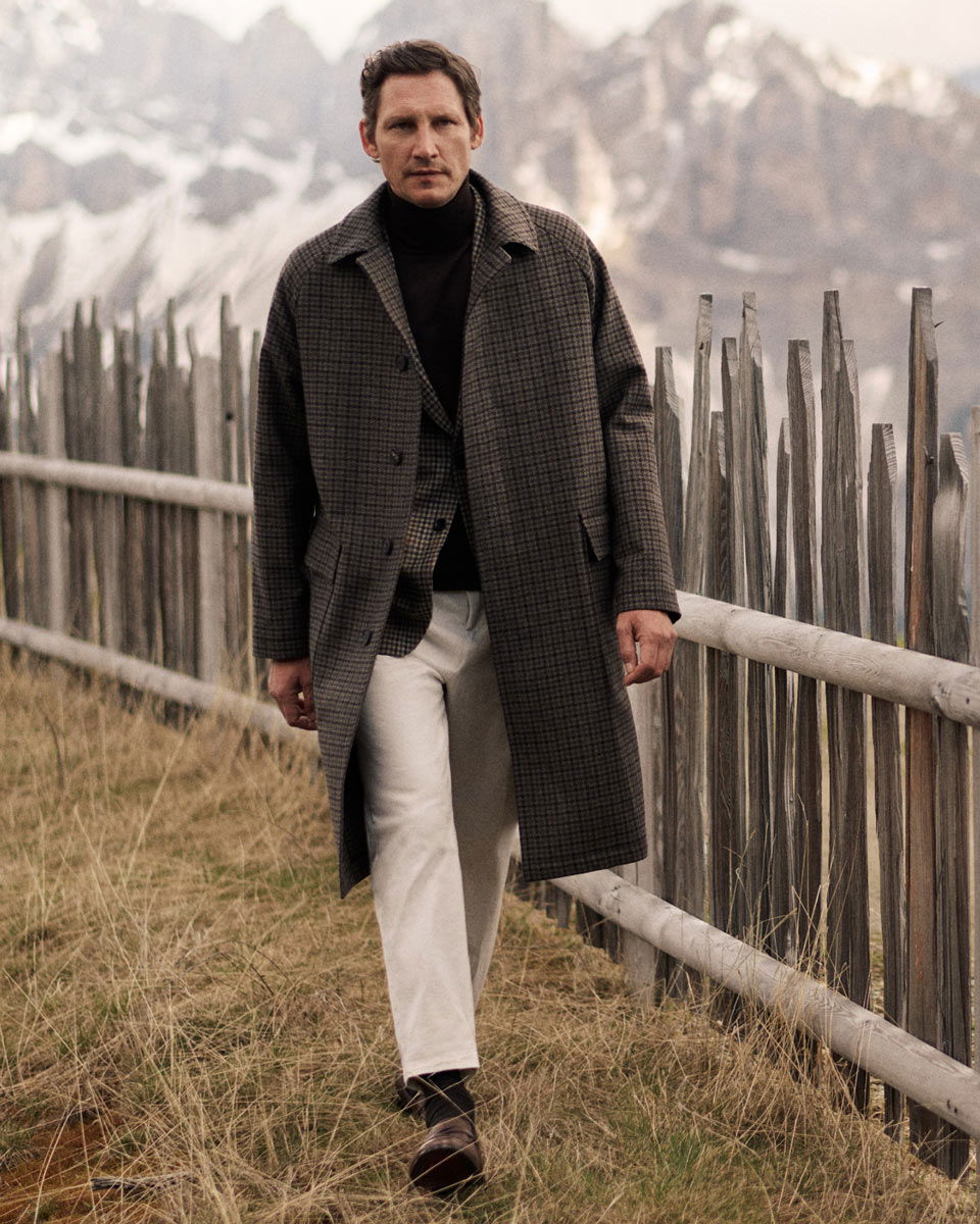 Mann trägt karierten Mantel, kariertes Sakko, schwarzen Rollkragenpullover und weiße Hose, geht am Holzzaun mit Bergblick. Mann trägt karierten Mantel, kariertes Sakko, schwarzen Rollkragenpullover und weiße Hose, geht am Holzzaun mit Bergblick.