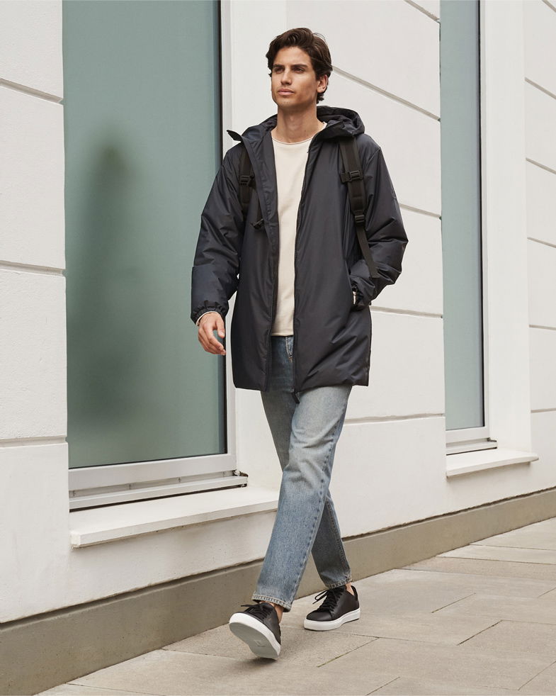Mann trägt beige Rundhalspullover, navy Kapuzen-Parka, graublaue Jeans, schwarzen Rucksack und navy Sneaker, geht auf Gehweg.