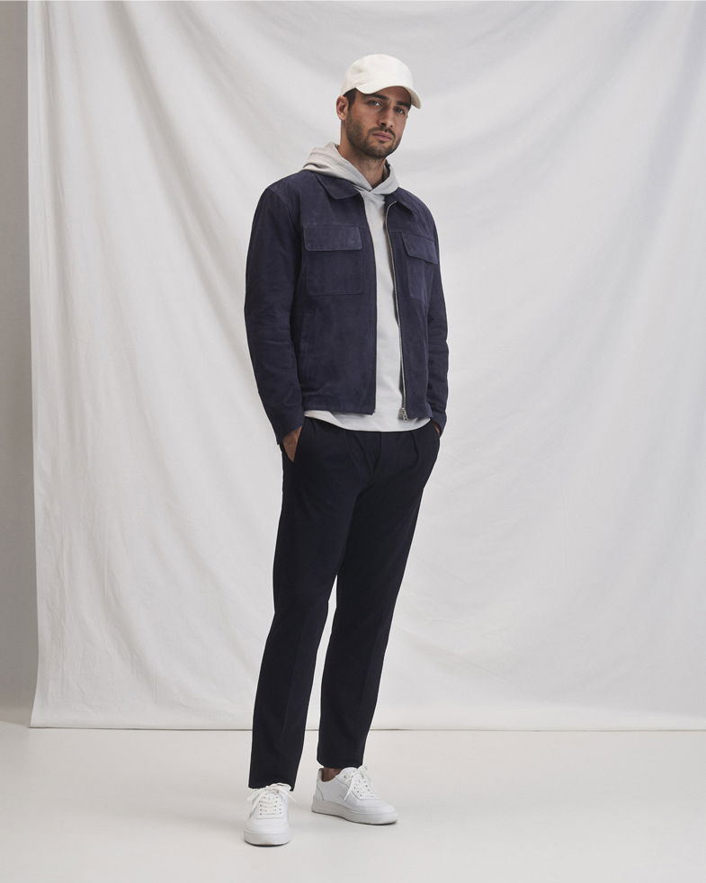 Mann trägt weiße Cap, grauen Hoodie, dunkelblaue Velourslederjacke, schwarze Hose und weiße Sneaker vor hellem Hintergrund.