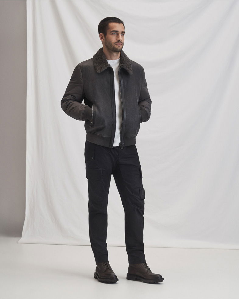 Mann trägt graue Lederjacke mit Fellkragen, weißes Shirt, schwarze Cargohose und braune Schnürboots vor weißem Hintergrund.