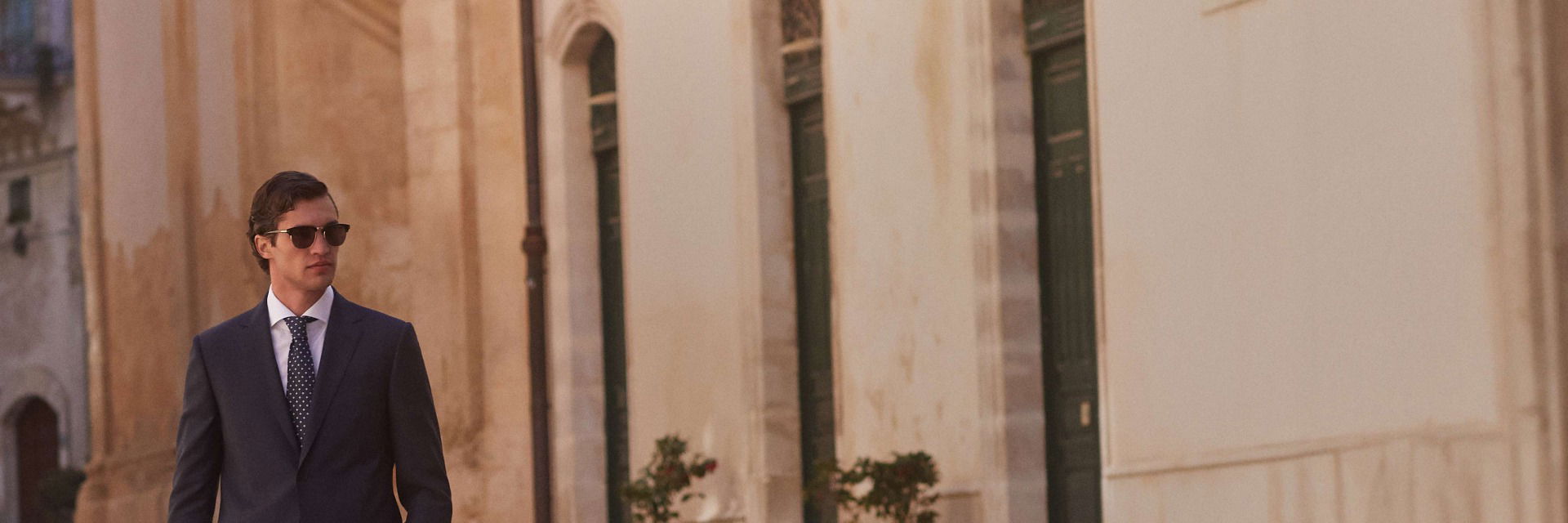 Mann in schwarzem Anzug, weißem Hemd, gepunkteter Krawatte und braunen Doppelmonk-Schuhen mit Sonnenbrille vor hellem Gebäude. Mann in schwarzem Anzug, weißem Hemd, gepunkteter Krawatte und braunen Doppelmonk-Schuhen mit Sonnenbrille vor hellem Gebäude.