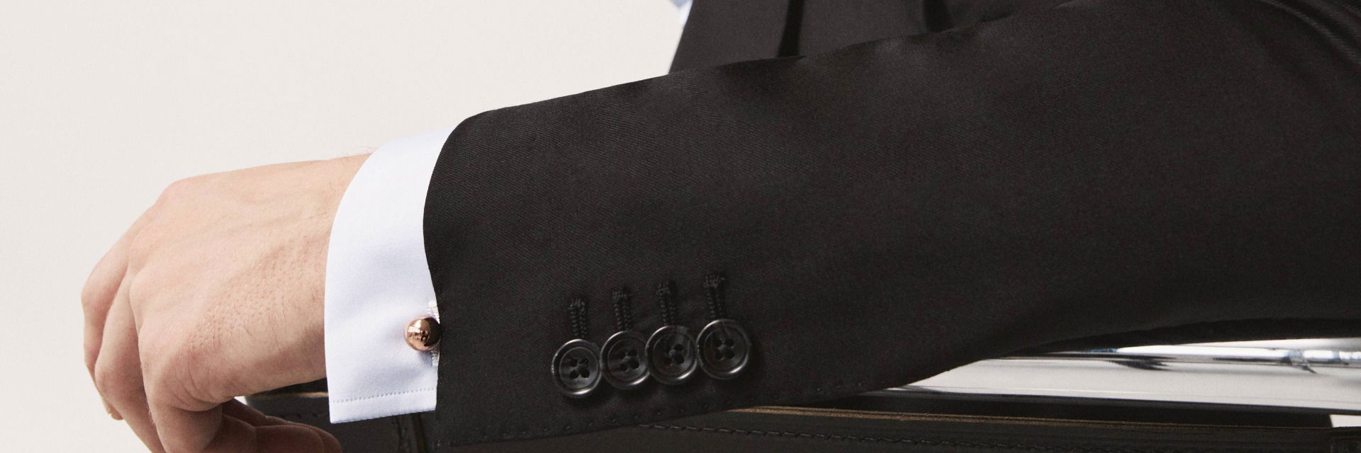 Schwarzes Sakko mit vier Knöpfen, weißes Hemd mit goldfarbenem runden Manschettenknopf an ausgestrecktem Arm. Schwarzes Sakko mit vier Knöpfen, weißes Hemd mit goldfarbenem runden Manschettenknopf an ausgestrecktem Arm.