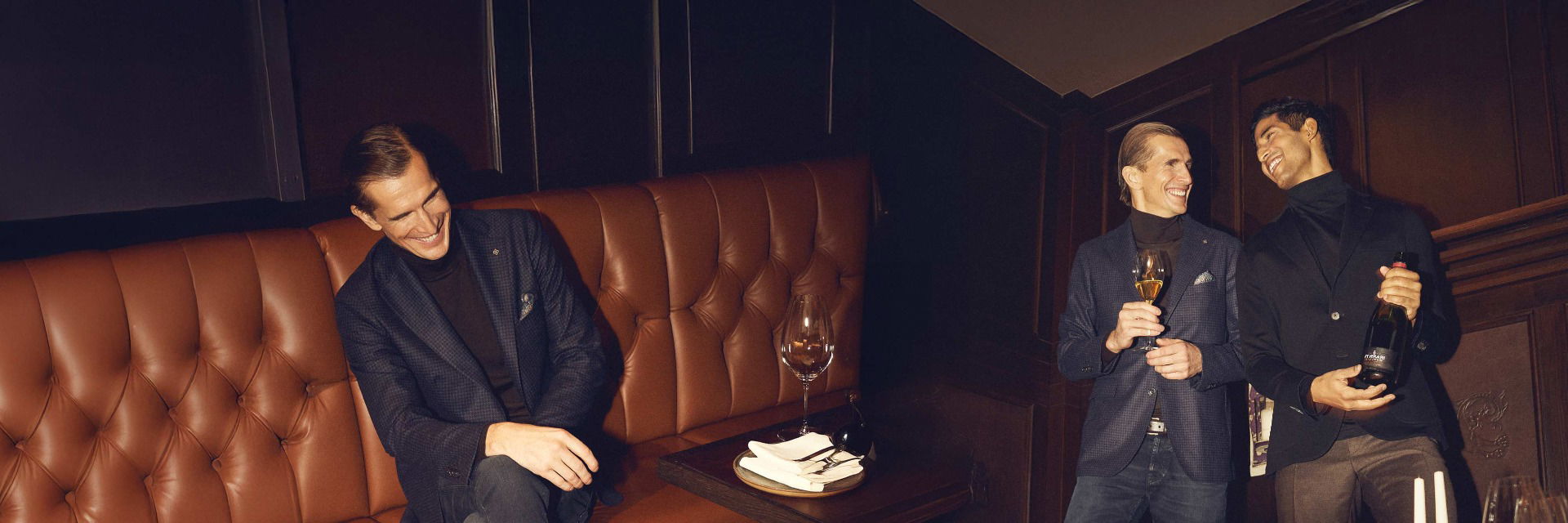 Drei Männer in Anzügen bei einer Feier, ein Mann sitzt lachend auf brauner Ledercouch, zwei stehen mit Getränken im eleganten Raum. Drei Männer in Anzügen bei einer Feier, ein Mann sitzt lachend auf brauner Ledercouch, zwei stehen mit Getränken im eleganten Raum.