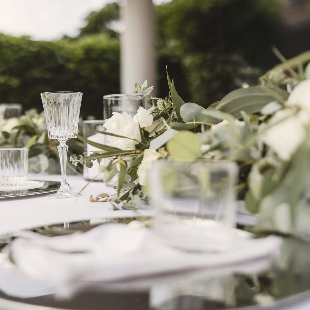 Eleganter gedeckter Hochzeitstisch im Freien mit Kristallgläsern, weißen Blüten und grünem Blumenschmuck.