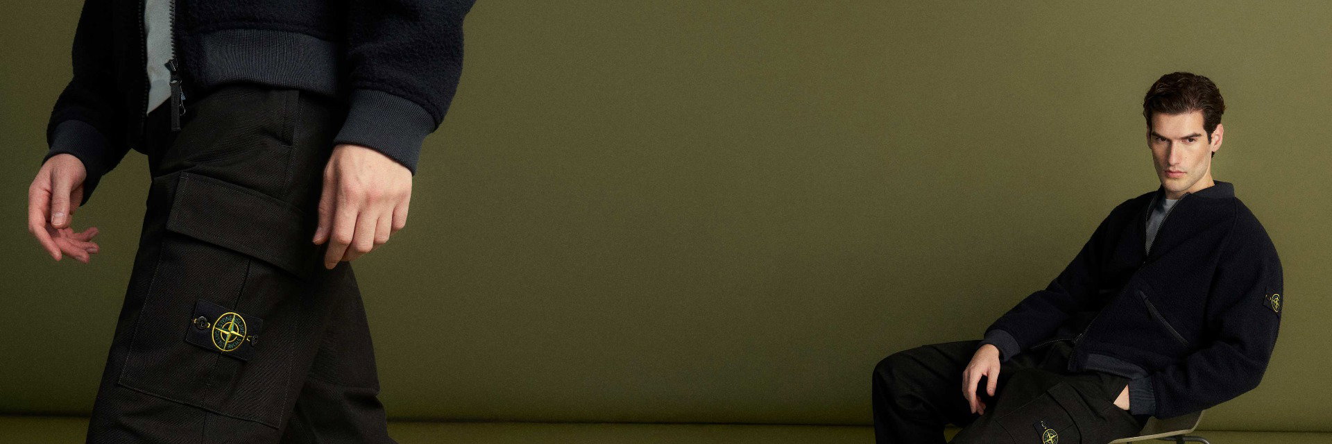 Nahaufnahme einer schwarzen Cargohose mit Taschen und Logo, daneben ein Mann in schwarzer Jacke sitzend vor grünem Hintergrund. Nahaufnahme einer schwarzen Cargohose mit Taschen und Logo, daneben ein Mann in schwarzer Jacke sitzend vor grünem Hintergrund.