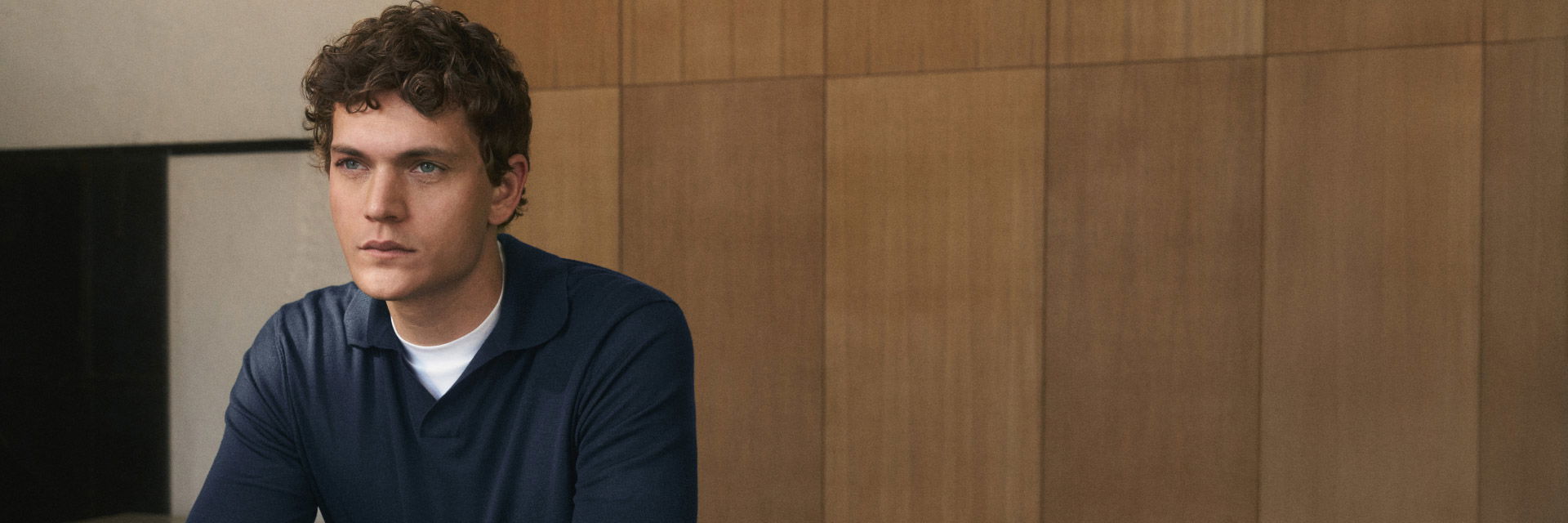 Junger Mann mit lockigem Haar trägt dunkelblaues Merino-Strickpolo über weißem T-Shirt vor Holzpaneelwand. Junger Mann mit lockigem Haar trägt dunkelblaues Merino-Strickpolo über weißem T-Shirt vor Holzpaneelwand.