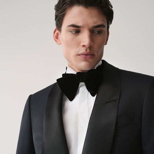 Junger Mann trägt schwarzen Smoking mit weißem Smokinghemd und schwarzer Fliege, blickt nach links, heller Hintergrund.