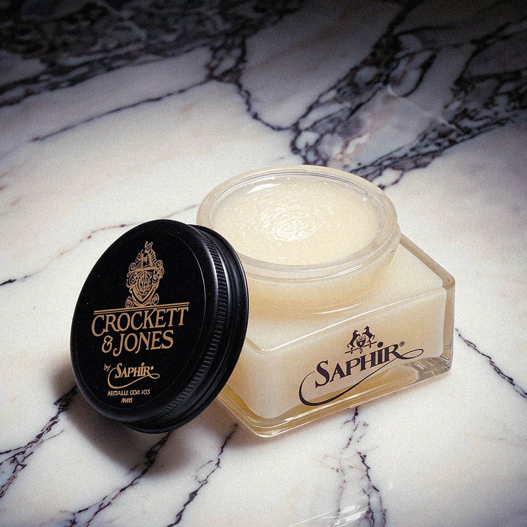 Offene Glasdose mit weißem Lederbalsam, schwarzer Deckel mit goldener Aufschrift 'Crockett & Jones by Saphir' auf Marmoruntergrund. Offene Glasdose mit weißem Lederbalsam, schwarzer Deckel mit goldener Aufschrift 'Crockett & Jones by Saphir' auf Marmoruntergrund.
