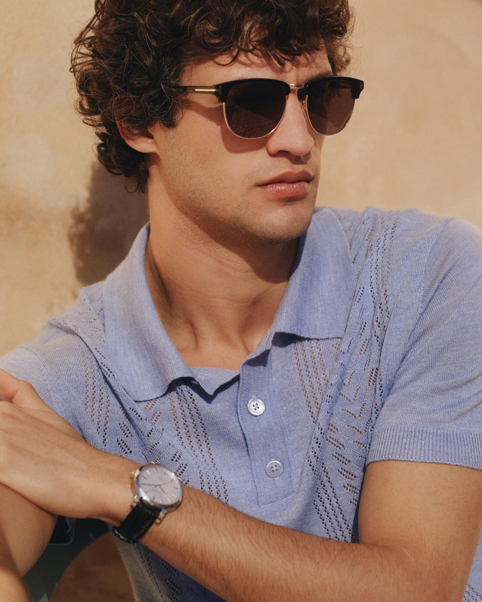 Junger Mann mit lockigem Haar, rauchblauem kurzarm Strickpolo, dunkler Sonnenbrille und silberner Armbanduhr mit schwarzem Lederarmband. Junger Mann mit lockigem Haar, rauchblauem kurzarm Strickpolo, dunkler Sonnenbrille und silberner Armbanduhr mit schwarzem Lederarmband.