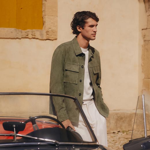 Mann mit olivgrünem Overshirt, weißem Kurzarmpullover und cremefarbener Hose lehnt an schwarzem Oldtimer-Cabrio vor gelber Wand. Mann mit olivgrünem Overshirt, weißem Kurzarmpullover und cremefarbener Hose lehnt an schwarzem Oldtimer-Cabrio vor gelber Wand.