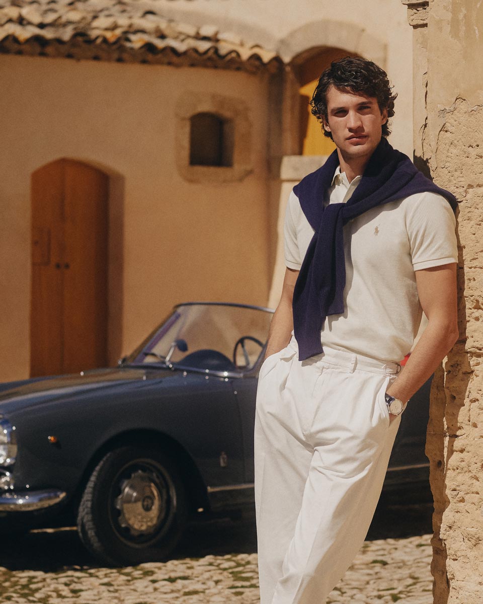 Junger Mann lehnt an Steinwand, trägt cremefarbenes Poloshirt, weiße Hose, navy Pullover um Schultern, Oldtimer Cabrio. Junger Mann lehnt an Steinwand, trägt cremefarbenes Poloshirt, weiße Hose, navy Pullover um Schultern, Oldtimer Cabrio.