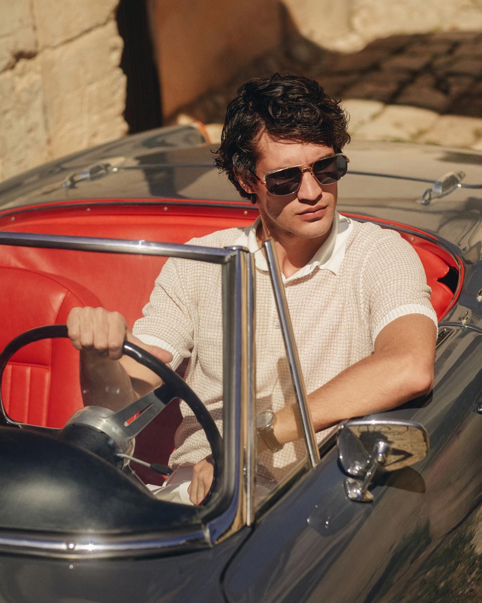 Junger Mann mit Sonnenbrille und beigem Kurzarm-Strickpolo sitzt in rotem Leder-Oldtimer-Cabriolet bei Sonnenschein. Junger Mann mit Sonnenbrille und beigem Kurzarm-Strickpolo sitzt in rotem Leder-Oldtimer-Cabriolet bei Sonnenschein.