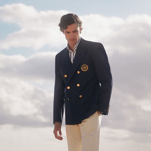 Junger Mann trägt navyblauen Zweireiher mit goldenen Knöpfen, blau-weiß gestreiftes Hemd und beige Hose vor wolkigem Himmel. Junger Mann trägt navyblauen Zweireiher mit goldenen Knöpfen, blau-weiß gestreiftes Hemd und beige Hose vor wolkigem Himmel.
