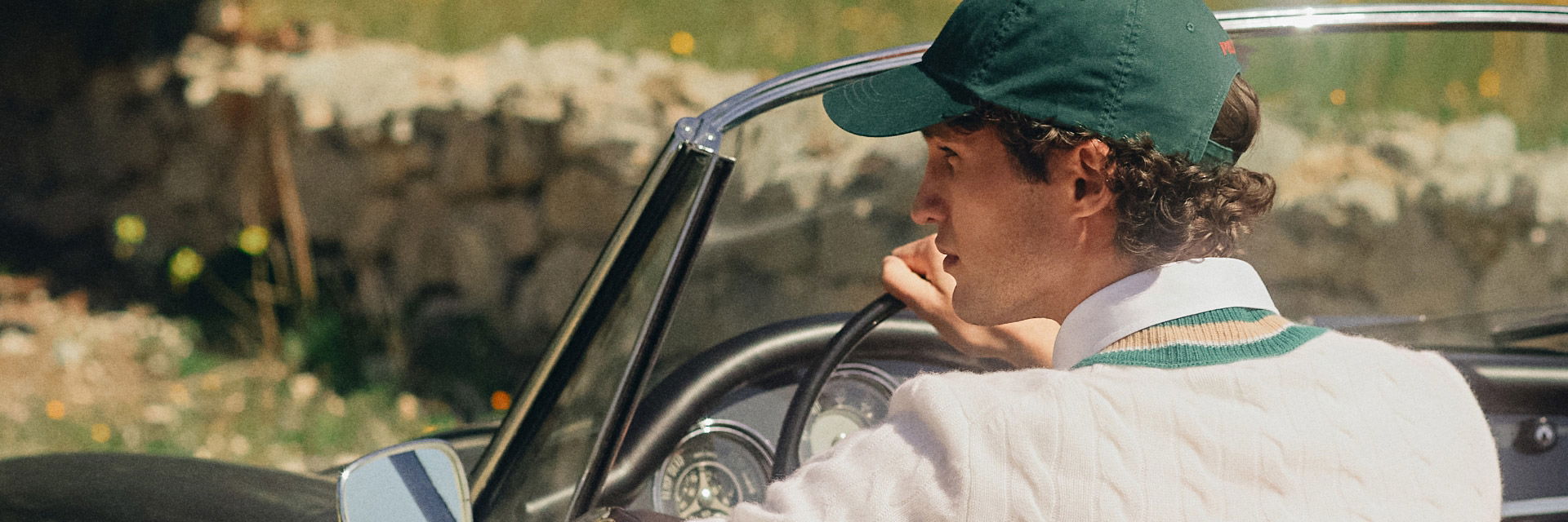 Junger Mann mit grünem Baseballcap und cremefarbenem V-Neck-Pullover fährt Oldtimer-Cabriolet bei Sonnenschein. Junger Mann mit grünem Baseballcap und cremefarbenem V-Neck-Pullover fährt Oldtimer-Cabriolet bei Sonnenschein.