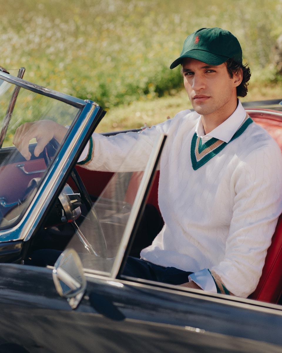 Junger Mann mit grünem Baseballcap und cremefarbenem V-Neck-Pullover sitzt im schwarzen Oldtimer-Cabriolet. Junger Mann mit grünem Baseballcap und cremefarbenem V-Neck-Pullover sitzt im schwarzen Oldtimer-Cabriolet.