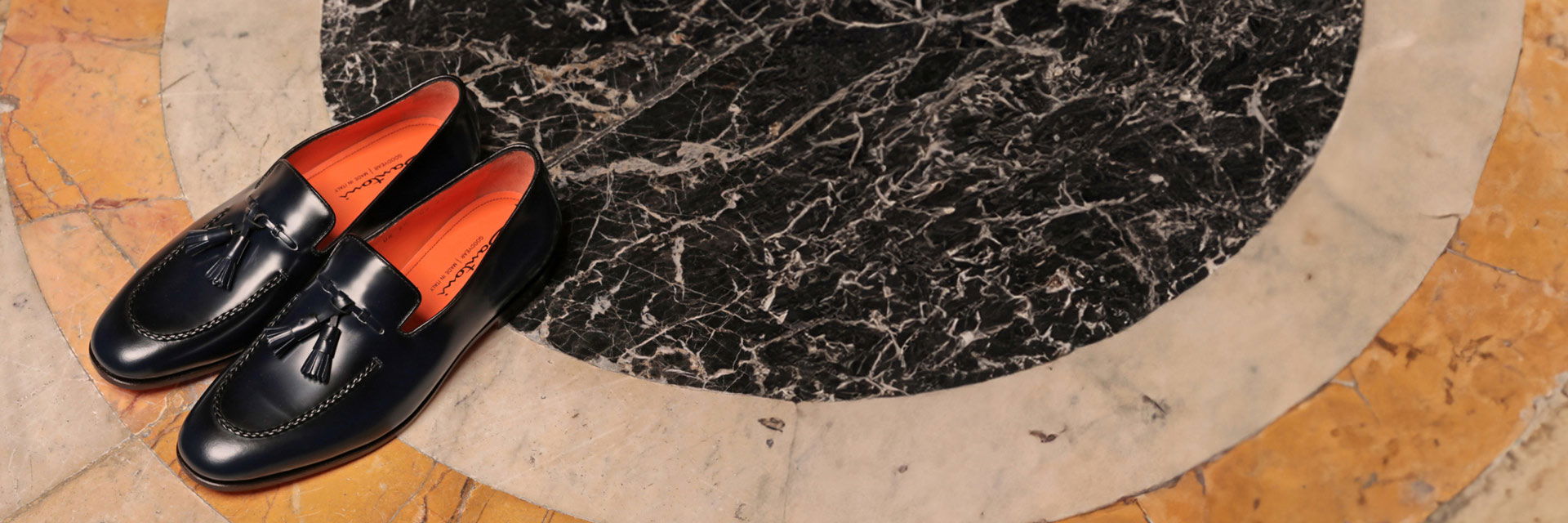 Schwarze Glattleder-Loafer für Herren mit Quasten, orange Innensohlen, auf rundem Marmorboden in Beige und Schwarz. Schwarze Glattleder-Loafer für Herren mit Quasten, orange Innensohlen, auf rundem Marmorboden in Beige und Schwarz.