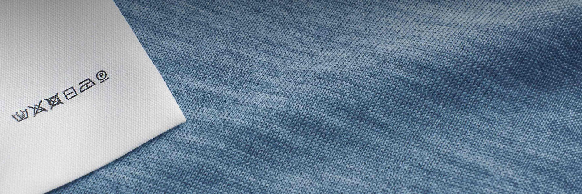 Pflegeetikett mit Waschsymbole für Jersey-Stoff in rauchblau, auf Stoffnaht genäht, Nahaufnahme der Textur. Pflegeetikett mit Waschsymbole für Jersey-Stoff in rauchblau, auf Stoffnaht genäht, Nahaufnahme der Textur.