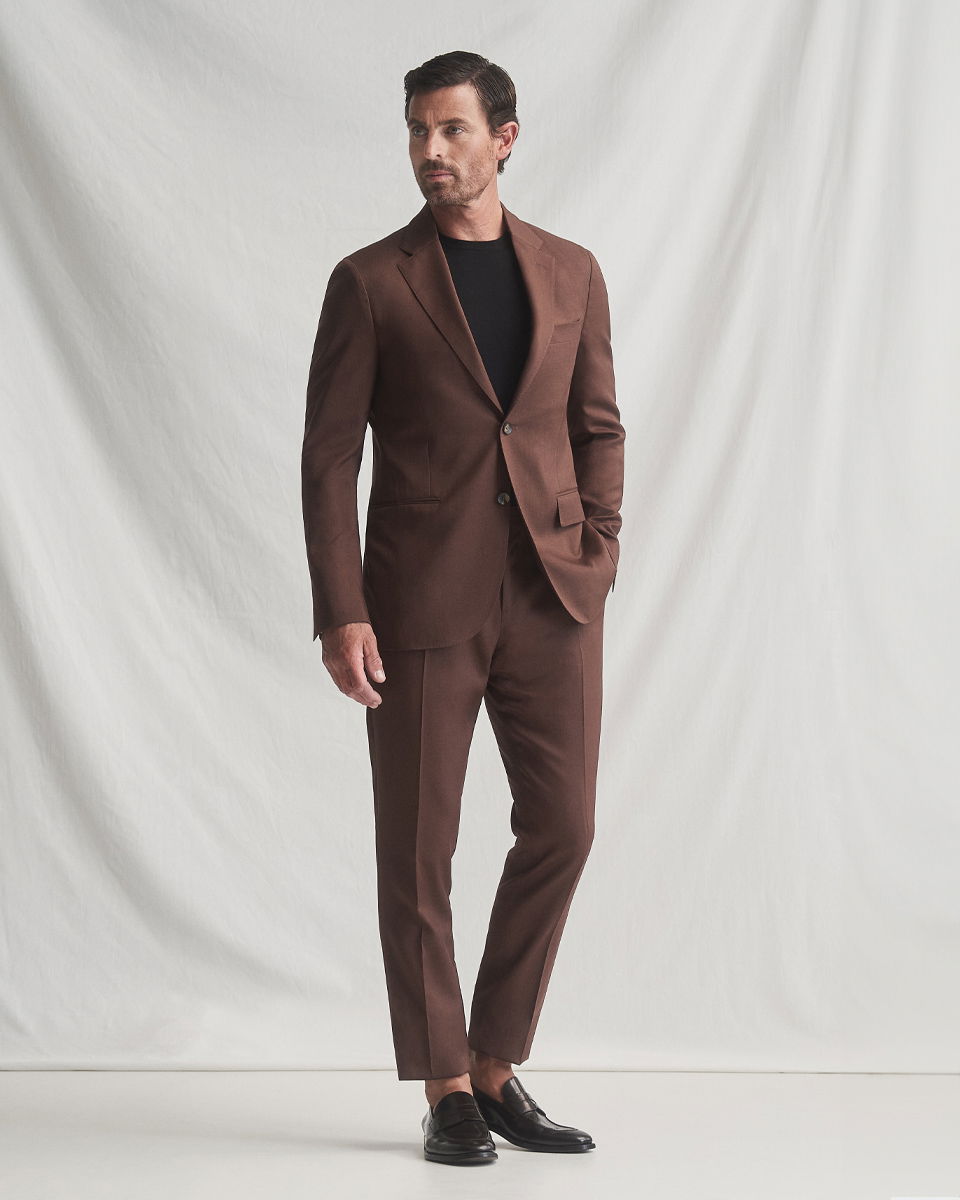 Mann trägt braunen Anzug mit schwarzem Rundhalsshirt und dunkelbraunen Lederloafern vor hellem Hintergrund, Hand in Hosentasche.