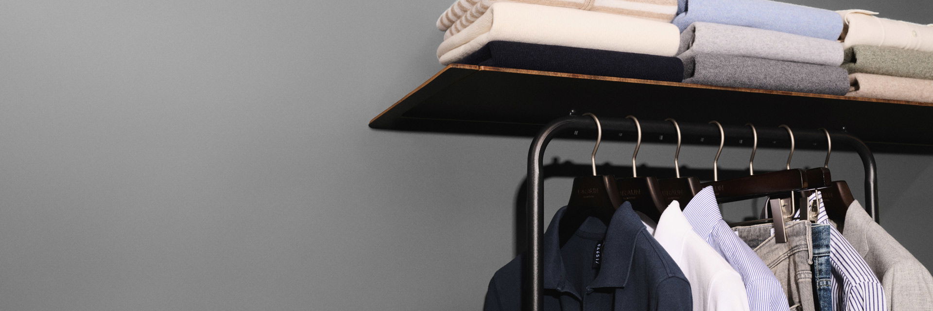 Regal mit gefalteten Pullovern in Pastellfarben, darunter Kleiderstange mit Hemden, Jeans und Sakkos auf Holzbügeln. Regal mit gefalteten Pullovern in Pastellfarben, darunter Kleiderstange mit Hemden, Jeans und Sakkos auf Holzbügeln.