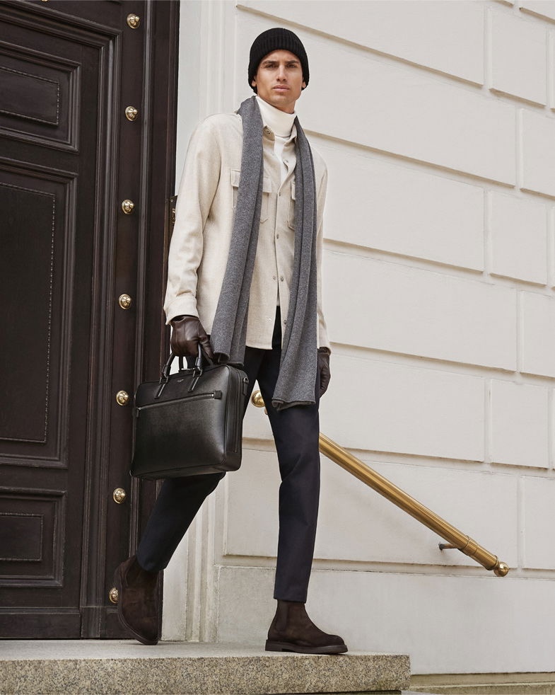 Mann trägt schwarzen Rollkragenpullover, beiges Overshirt, schwarze Hose, braune Boots, grauen Schal, schwarze Mütze und hält schwarze Tasche.