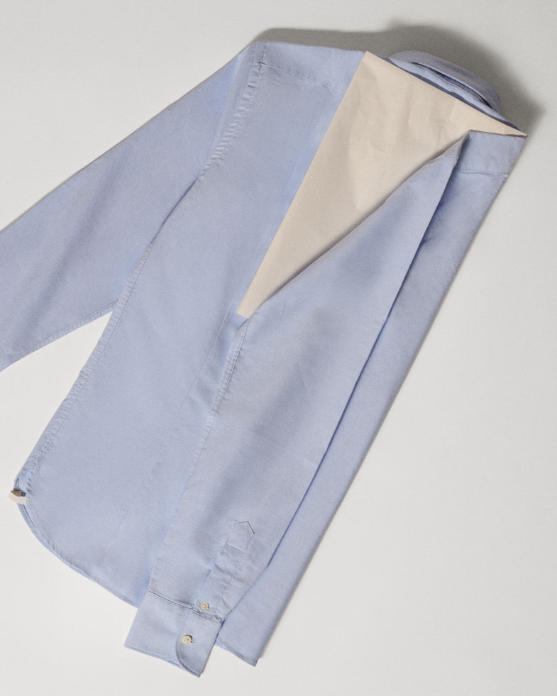 Hellblaues Herrenhemd, zur Hälfte gefaltet, Rückansicht, auf hellem Untergrund liegend, mit sichtbarem Kragen und Manschetten.