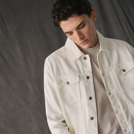 Junger Mann mit dunklen Haaren, cremefarbenem Shirt und weißer Jeansjacke vor grauem Hintergrund, schaut nach unten.