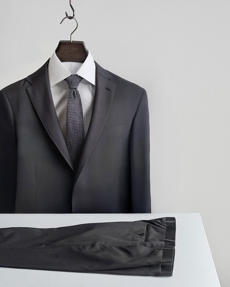 Anthrazitfarbenes Sakko mit weißem Hemd und grauer Krawatte auf Bügel, schwarze Hose liegt auf weißem Tisch. Anthrazitfarbenes Sakko mit weißem Hemd und grauer Krawatte auf Bügel, schwarze Hose liegt auf weißem Tisch.
