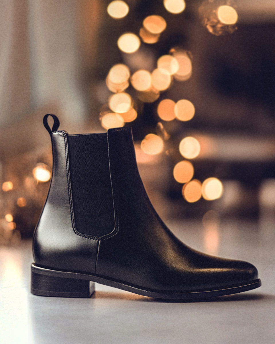 Schwarzer Chelsea-Boot für Herren in Seitenansicht, mit elastischem Einsatz und Lichter-Dekoration im unscharfen Hintergrund. Schwarzer Chelsea-Boot für Herren in Seitenansicht, mit elastischem Einsatz und Lichter-Dekoration im unscharfen Hintergrund.