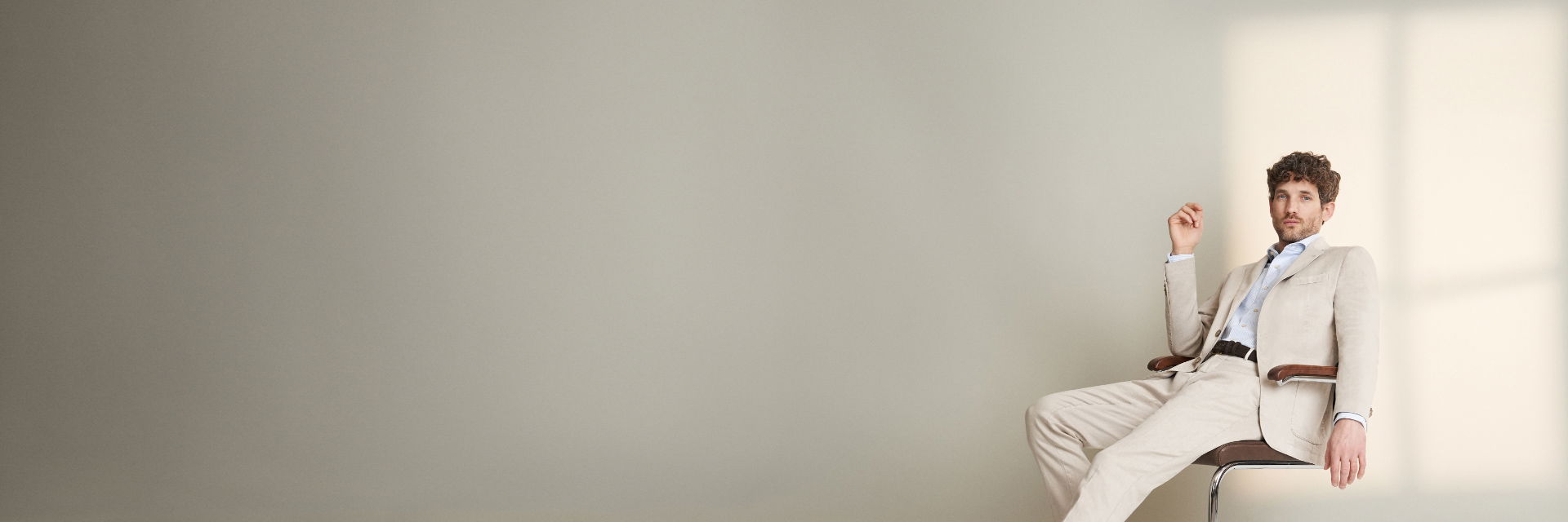 Sitzender Mann im beigen Anzug und hellblau gestreiftem Hemd vor heller Wand, entspannte Pose auf Stuhl. Sitzender Mann im beigen Anzug und hellblau gestreiftem Hemd vor heller Wand, entspannte Pose auf Stuhl.